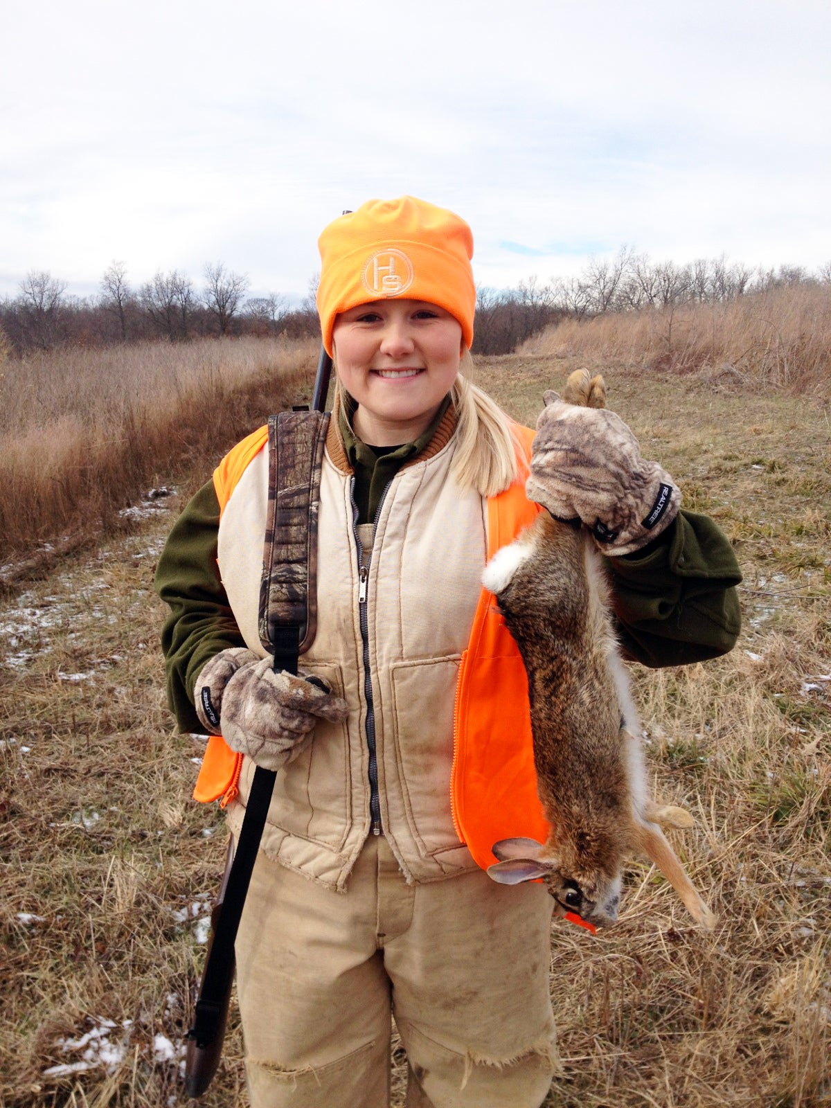 A warmly-dressed girl with a rifle slung over her shoulder holds up a rabbit she harvested.