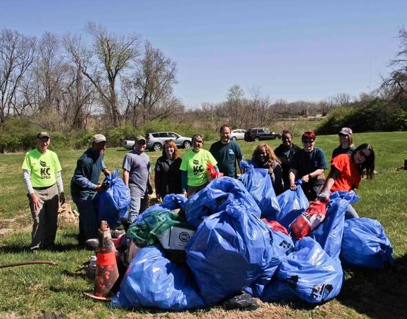 volunteers pose before a large pile of blue trash bags full of garbage retrieved from a river.