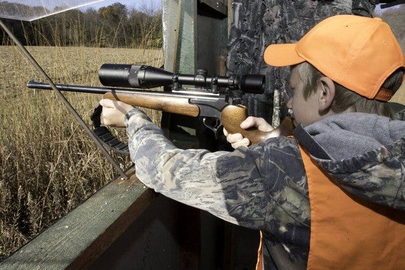 Young deer hunter aims rifle in deer blind