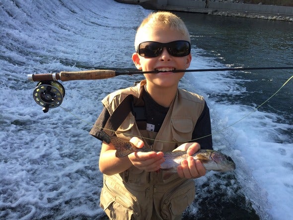 Boy holds rainbow trout while holding fishing rod in mouth at Bennett Spring State Park.