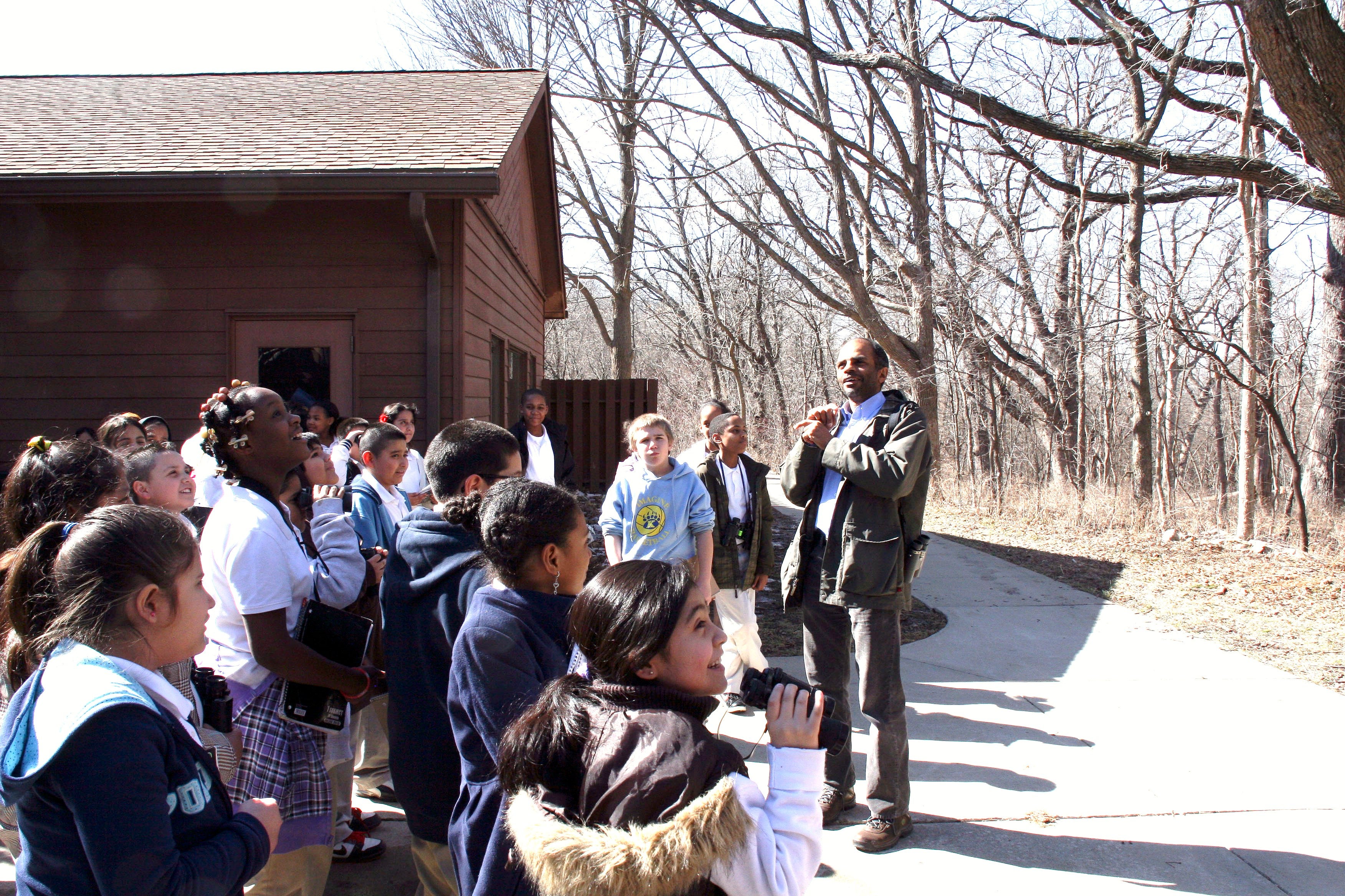 An ornithologist instructs a group of schoolchildren how to birdwatch. Several of the children are holding binoculars.