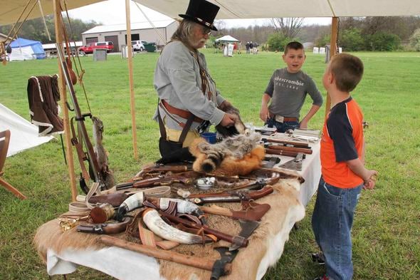 A man in frontier garb shows a table full of pelts to two boys. 