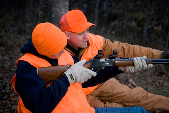 A mentor helps a hunter aim a rifle