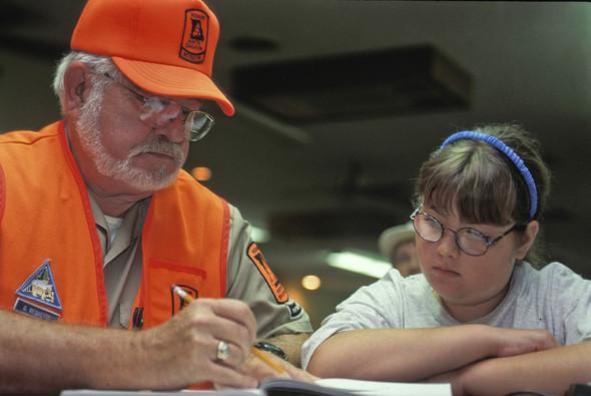 A girl reading hunter education materials. She has a shotgun and a safety vest in front of her.