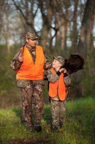Father and son wearing hunter orange return from a successful turkey hunt. The boy has a turkey over his shoulder.
