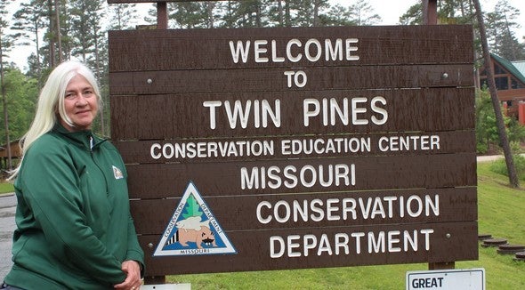 Reva Dow stands next to the entrance sign to Twin Pines Conservation Center