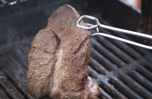 A pair of tongs flips a venison steak on a grill.