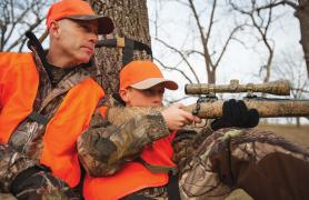 An adult mentor sits behind a young hunter with a rifle. 