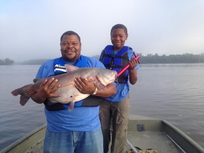 Father and son hold catfish on a boat.