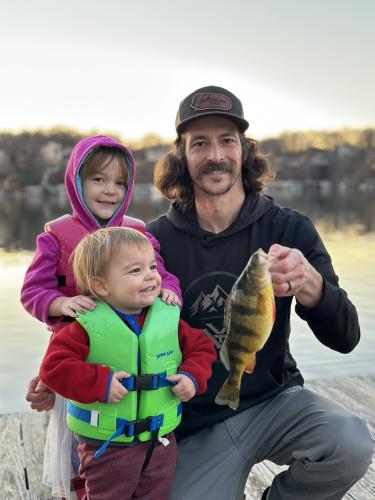 Jeffrey Needles holding a yellow perch with his two children standing next to him.