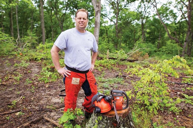 A man wearing orange safety chaps stands by a tree stump. His chainsaw, gloves, helmet, and visor are on the stump. 