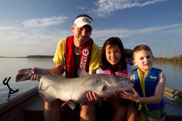 An adult angler and two children hold a large catfish in their boat