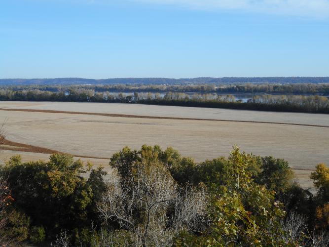 A view of fields down a hill from an overlook. The Mississippi River flows in the distance behind a row of trees.