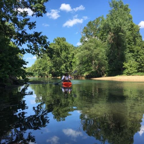 A person in a hat is paddling a kayak along a tree-lined stream. The blue sky, clouds, and trees are reflected in the water.