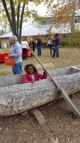a young girl sits in a dugout canoe and is holding up a heavy paddle in front of the Discovery Center. 