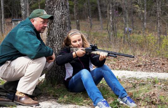 An adult mentor watches as a girl take aim at a turkey
