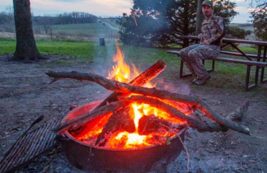 A man in camoflage hunting gear sits at a picnic table by a campfire in a fire ring.