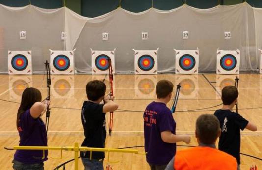 Students lined up to shoot at archery targets in a gym.