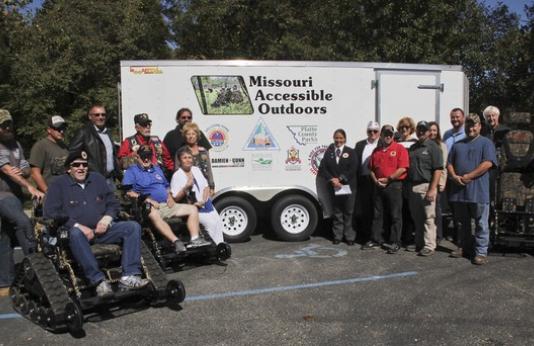 A group of people present three track chairs to MDC. The trailer that transports the chairs is in the background.